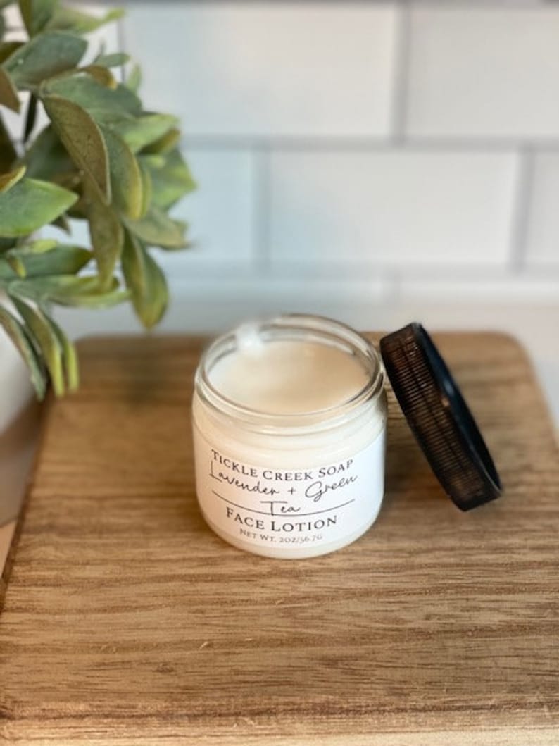 May include: A white jar of face lotion with a black lid. The label reads "Tickle Creek Soap Lavender + Green Tea Face Lotion Net Wt 2oz/56.7g".