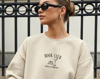 Puede incluir: Sudadera beige con el texto "BOOK CLUB" y un pequeño gráfico. La sudadera se combina con botas marrones, un bolso beige y gafas de sol. La persona está sentada en una acera con hojas de otoño.