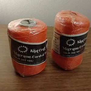 May include: Two spools of bright orange Knot Craft macrame cord and twine, each wrapped in clear plastic. The black labels feature a sun logo and text stating 306 ft. (102 yds.) of cord. The spools are cylindrical with a hollow center core.