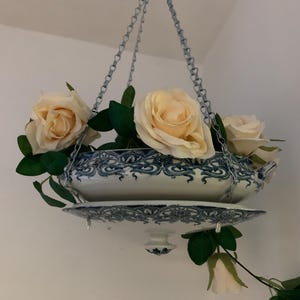 May include: A white porcelain planter with blue floral designs hangs from a silver chain. The planter is filled with artificial white roses and green leaves.