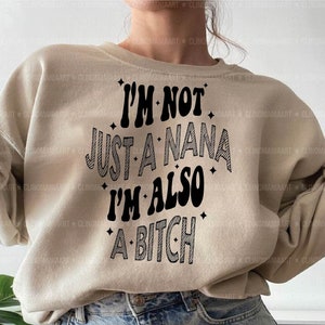 以下が含まれることがあります： 黒色の「I'M NOT JUST A NANA I'M ALSO A BITCH」というテキストが入ったベージュ色のスウェットシャツ。単語は太字で遊び心のあるフォントです。画像には「Digital Download SVG | PNG | PDF」という単語も含まれています。