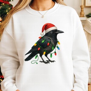 Può includere: Felpa bianca con un'illustrazione di un corvo nero con un cappello di Babbo Natale e luci natalizie. Il corvo è decorato con luci colorate. Il testo "Digital Download | PNG" è in basso.
