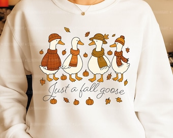 May include: White sweatshirt featuring a fall-themed graphic. The design includes four cartoon geese wearing hats and scarves, surrounded by pumpkins and leaves. The text "Just a fall goose" is also present.