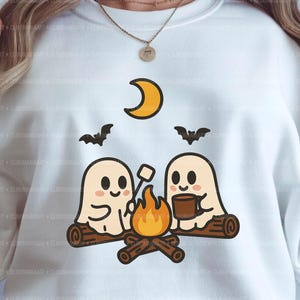 Peut inclure: Sweat-shirt blanc avec un motif sur le thème d'Halloween. Le motif comprend deux fantômes de dessins animés grillant des guimauves autour d'un feu de camp, avec un croissant de lune et des chauves-souris. Le texte en bas indique "Digital Download | PNG".