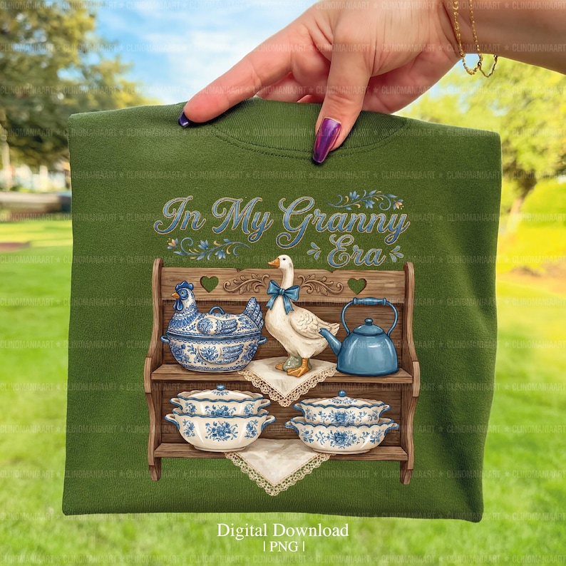 May include: Green t-shirt featuring a graphic of a wooden shelf with a chicken-shaped dish, a goose with a blue bow, and a blue teapot. The shelf also holds blue and white floral dishes. The text "In My Granny Era" is above the shelf.