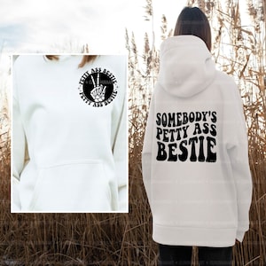 Op de afbeelding: Witte hoodie met een zwarte grafische print op de rug die "Somebody's Petty Ass Bestie" zegt. De voorkant van de hoodie heeft een kleinere zwarte grafische print die "Petty Ass Bestie" zegt met een vredesteken.