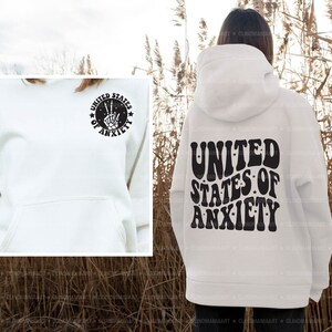 Peut inclure: Sweat-shirt &agrave; capuche blanc avec un motif noir au dos qui dit "United States of Anxiety" dans une police r&eacute;tro. Le motif comprend &eacute;galement de petites &eacute;toiles. Le devant du sweat-shirt a un plus petit motif noir d'un signe de paix avec une main &agrave; l'int&eacute;rieur d'un cercle avec le texte "United States of Anxiety".