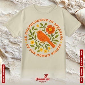 May include: Cream-colored t-shirt featuring a circular design with an orange bird, flowers, and the text "BE UNAPOLOGETIC IN SPEAKING UP FOR HUMAN RIGHTS." The design includes green leaves and yellow flowers. The shirt has short sleeves.