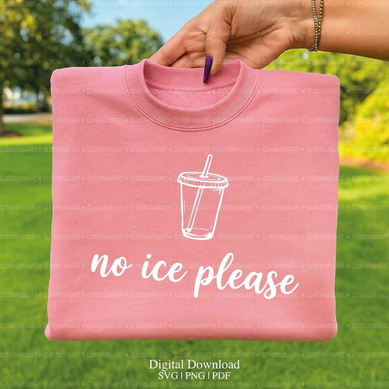 May include: A light pink sweatshirt with a white graphic of a drink cup and the words "no ice please". The sweatshirt is folded and held up by a hand against a green background.