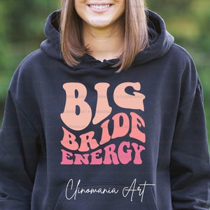 K&ouml;nnte beinhalten: Schwarzer Hoodie mit einem pinkfarbenen und orangenen Aufdruck, der "Big Bride Energy" und "Cinomania Art" in wei&szlig;er Schrift zeigt.