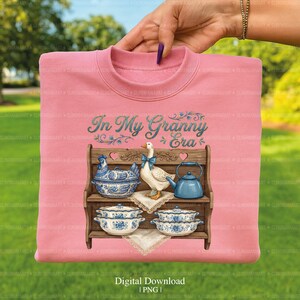 May include: Pink sweatshirt with the text "In My Granny Era" in teal script. The design features a wooden shelf with blue and white ceramic dishes, a goose with a blue bow, and a blue teapot.