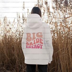 K&ouml;nnte beinhalten: Wei&szlig;er Hoodie mit einem pinkfarbenen und orangenen Aufdruck, der auf der R&uuml;ckseite "Big Bride Energy" lautet.