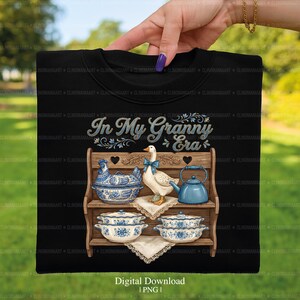 May include: Black t-shirt featuring a graphic of a wooden shelf with blue and white porcelain dishes, a goose with a blue bow, and a teapot. The text "In My Granny Era" is above the shelf. Digital download.