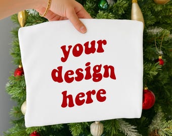 Könnte beinhalten: Ein weißer Kissenbezug mit dem roten Schriftzug "your design here" wird vor einem Weihnachtsbaum gehalten. Der Baum ist mit roten, goldenen und grünen Kugeln geschmückt. Ein goldenes Armband ist sichtbar.