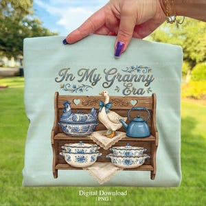 May include: A light blue t-shirt featuring a vintage-style illustration. The design includes a wooden shelf with blue and white ceramic dishes, a goose with a blue bow, and the text "In My Granny Era".