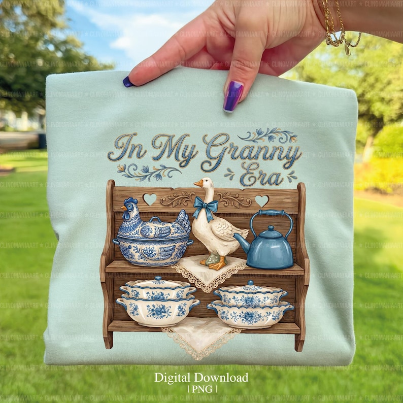 May include: A light blue t-shirt featuring a vintage-style illustration. The design includes a wooden shelf with blue and white ceramic dishes, a goose with a blue bow, and the text "In My Granny Era".