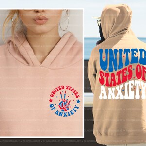 Peut inclure: Un sweat-shirt rose et beige avec un graphique rouge, blanc et bleu qui dit "United States of Anxiety" avec un signe de paix et des &eacute;toiles.