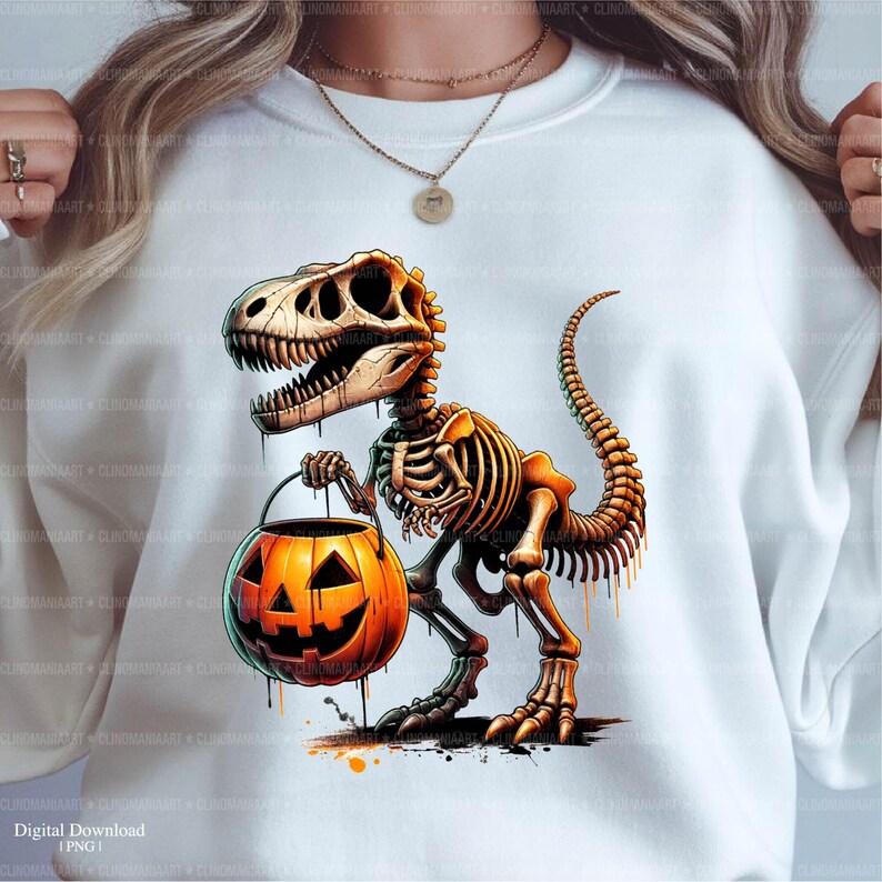 May include: White sweatshirt featuring a Halloween-themed graphic. The design showcases a dinosaur skeleton holding an orange jack-o'-lantern. The dinosaur is brown and beige, with a detailed skeletal structure. The pumpkin has a carved face.