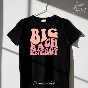 May include: Black t-shirt featuring a pink and peach graphic that reads "BIG BACH ENERGY" in a retro font. The shirt is hanging on a hanger against a white wall.