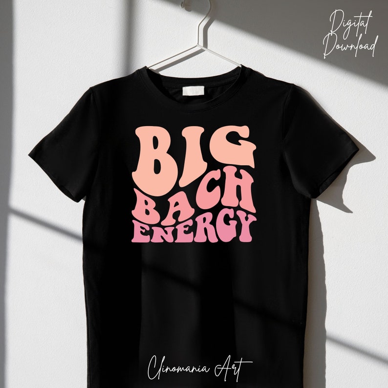 May include: Black t-shirt featuring a pink and peach graphic that reads "BIG BACH ENERGY" in a retro font. The shirt is hanging on a hanger against a white wall.