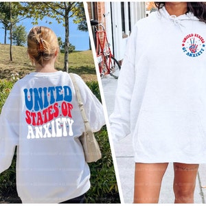 Peut inclure: Deux sweat-shirts blancs avec un motif graphique rouge, blanc et bleu. Un sweat-shirt porte le texte "United States of Anxiety" dans une police r&eacute;tro. L'autre sweat-shirt a un signe de paix avec des &eacute;toiles et le texte "United States of Anxiety".