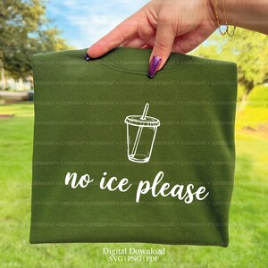May include: An olive green sweatshirt with a white graphic of a beverage cup and the words "no ice please". The sweatshirt is held up against a blurred outdoor background.