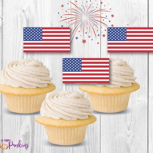 Puede incluir: Tres cupcakes de vainilla con glaseado blanco, decorados con pequeños adornos de bandera estadounidense. Las banderas son rojas, blancas y azules con estrellas. Los cupcakes están sobre un fondo de madera blanca.