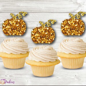 May include: Three cupcakes with light yellow cake and swirled beige frosting, topped with glittery gold pumpkin decorations. The pumpkins have curly stems. The cupcakes are arranged in a row. The logo "Lil Purple Punkins" is in the bottom left corner.