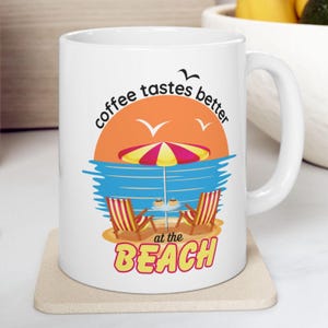 May include: White ceramic coffee mug with a beach scene and the text "coffee tastes better at the beach". The scene features a beach umbrella, two beach chairs, and two cups of coffee on a small table.