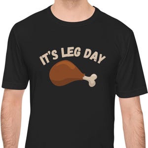 May include: Black t-shirt with the phrase "IT'S LEG DAY" in white and a cartoon image of a roasted chicken leg in brown and white. The shirt is short-sleeved.