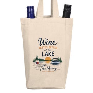 May include: A beige canvas wine tote bag with two bottles inside. The bag features the text "Wine tastes better at the Lake" with a lake scene illustration and "Lake Murray South Carolina" text.