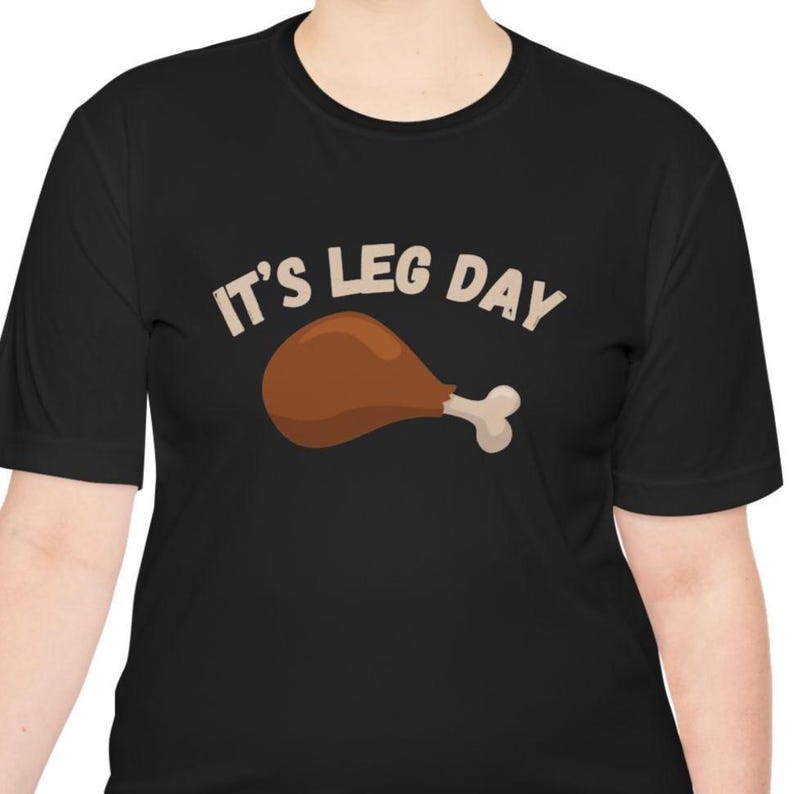 May include: Black t-shirt with the text "IT'S LEG DAY" above a cartoon image of a roasted turkey leg. The turkey leg is brown with a white bone. The text and graphic are in a light tan color.