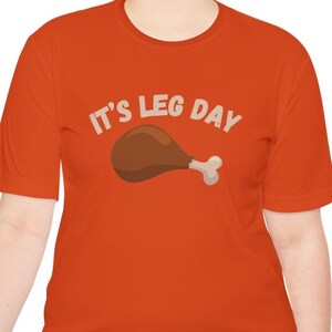 May include: Orange t-shirt with the text "IT'S LEG DAY" in white letters above a cartoon image of a roasted turkey leg. The shirt is a solid orange color.