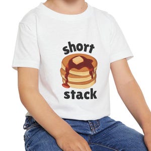 Può includere: Maglietta bianca con una grafica di un piccolo stack di pancake con sciroppo e burro. Il testo "short stack" è stampato sotto i pancake.