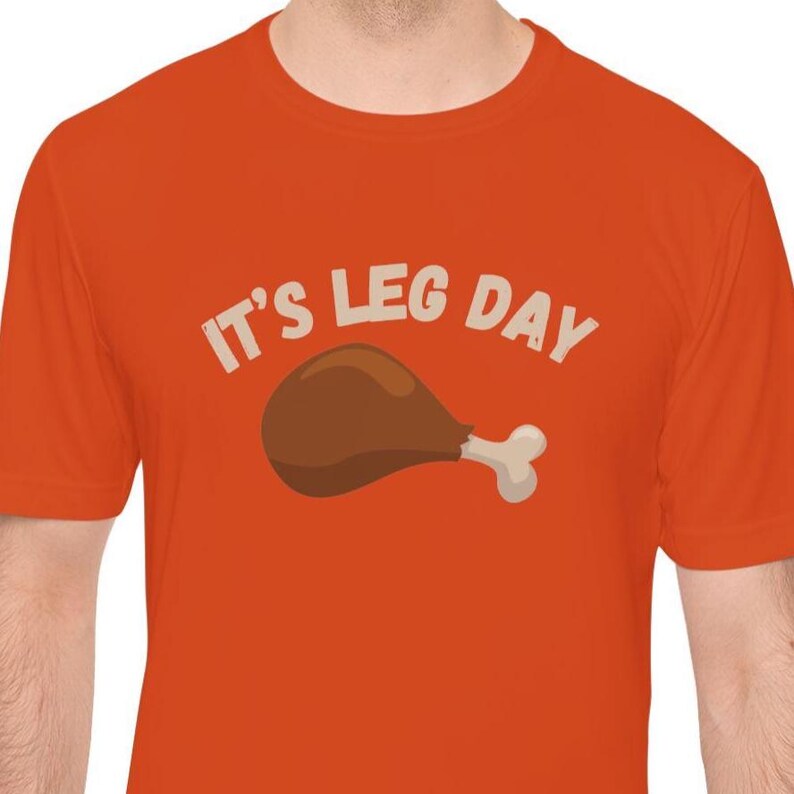 May include: Orange t-shirt with the text "IT'S LEG DAY" in white and a cartoon image of a cooked chicken leg in brown and white. The shirt is short-sleeved and has a crew neck.