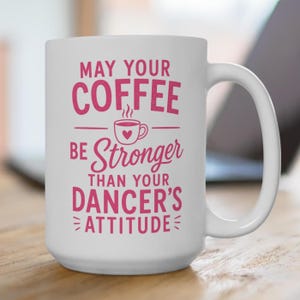 May include: A white ceramic mug featuring pink text that reads, "MAY YOUR COFFEE BE STRONGER THAN YOUR DANCER'S ATTITUDE," with a small graphic of a coffee cup containing a heart in the center, positioned between the lines of text.