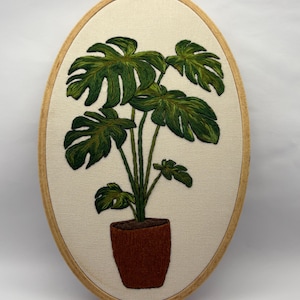 May include: An embroidered artwork featuring a green monstera plant in a brown pot, set against a cream-colored fabric. The oval frame is made of light wood. The leaves have detailed shading, and the pot has a textured appearance.