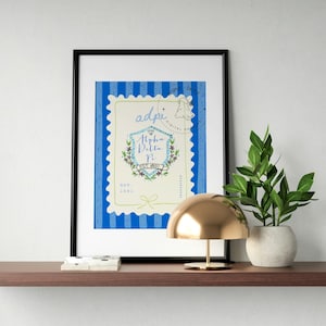 May include: A framed print with a blue and white design, displaying the text "adpi Alpha Delta Pi Est. 1851." The print is on a brown shelf, alongside a gold lamp and a small potted plant.