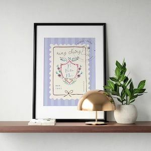 May include: Framed art print with a postage stamp design, featuring the words "ring ching" and "Pi Beta Phi." The artwork includes floral and wing illustrations on a lavender striped background. A gold lamp and a potted plant sit on a wooden shelf.
