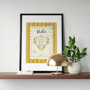 Puede incluir: Una impresión enmarcada con un diseño amarillo y blanco, el texto "theta" y "Kappa Alpha Theta". La impresión está en un marco negro y se exhibe en un estante marrón con una lámpara dorada y una planta en maceta.