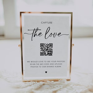 May include: A gold-framed sign with a white background and black text that reads "CAPTURE the love". A black and white QR code is below the text. The sign also includes the text "WE WOULD LOVE TO SEE YOUR PHOTOS! SCAN THE QR CODE AND UPLOAD PHOTOS TO OUR SHARED ALBUM."