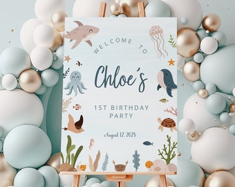 May include: A blue and white welcome sign with a sea-themed design. The sign says "Welcome to Chloe's 1st Birthday Party" with a date of August 12, 2023. The sign features illustrations of an octopus, a whale, a turtle, a shark, and other sea creatures.
