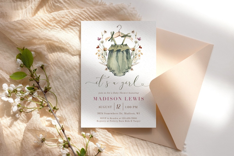 May include: It's a girl baby shower invitation with a watercolor illustration of a green onesie with a floral wreath. The invitation includes the text "It's a girl", "Madison Lewis", "August 12", "1:00 PM", "5674 Somewhere Dr. Madison, WI", "RSVP to Kendra at 123-456-7890", and "Registry at Pottery Barn Kids & Target".