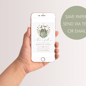 May include: A digital baby shower invitation on a smartphone screen. The invitation features a watercolor illustration of a baby onesie with flowers and the text "it's a girl" in pink and white. The invitation also includes the name "MADISON LEWIS" and the date and time of the shower.