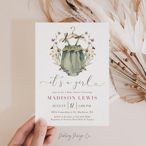 May include: It's a girl baby shower invitation with a watercolor illustration of a green baby onesie with a floral wreath. The invitation includes the text "It's a girl", "Join us for a Baby Shower honoring Madison Lewis", "August 12 | 1:00 PM", "5674 Somewhere Dr, Madison, WI", "RSVP to Kendra at 123-456-7890", "Registry at Pottery Barn Kids & Target", and "Dahling Design Co."