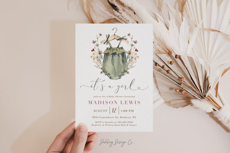 May include: It's a girl baby shower invitation with a watercolor illustration of a green baby onesie with a floral wreath. The invitation includes the text "It's a girl", "Join us for a Baby Shower honoring Madison Lewis", "August 12 | 1:00 PM", "5674 Somewhere Dr, Madison, WI", "RSVP to Kendra at 123-456-7890", "Registry at Pottery Barn Kids & Target", and "Dahling Design Co."