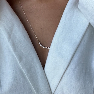 Puede incluir: Un delicado collar de oro con una fila de peque&ntilde;as piedras preciosas transparentes. El collar lo lleva una persona que lleva una chaqueta de lino blanca.