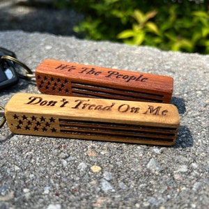 May include: Two wooden keychains with American flag designs. The top keychain says "We The People" and the bottom keychain says "Don't Tread On Me".