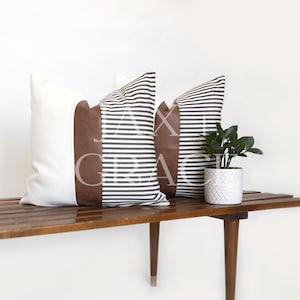 May include: Two decorative throw pillows with a brown leather accent and white and black striped fabric. The pillows are sitting on a wooden bench.