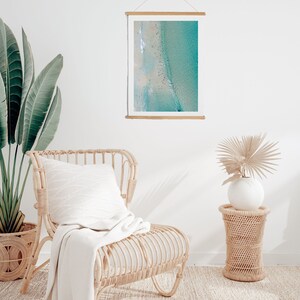 May include: A framed print of a turquoise ocean scene hangs on a white wall. A woven rattan chair with a white pillow and blanket sits nearby. A potted plant and a small woven side table with a vase and dried plant complete the coastal decor.